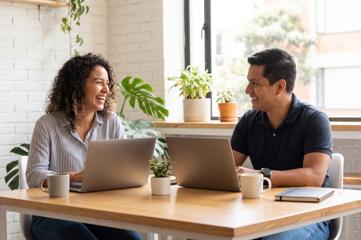 Two colleagues laughing together at their laptops at a sunny shared table